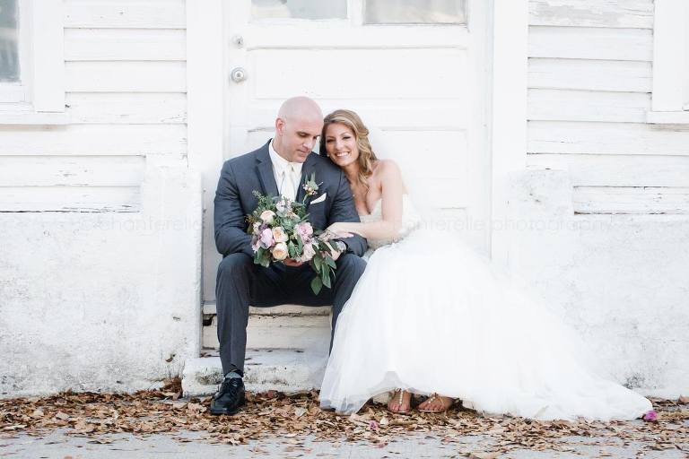 bride and groom sitting against white rustic house